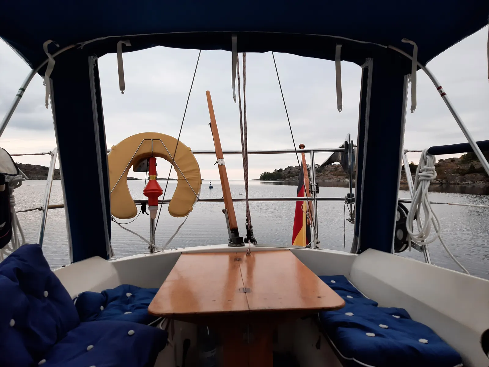 Vy inifrån ett mörkblått sittbrunnskapell: blå knappade dynor på båda sidor om ett fällbart träbord, och genom öppningen en livboj, stilla vatten och klippiga skär i kvällsljus.