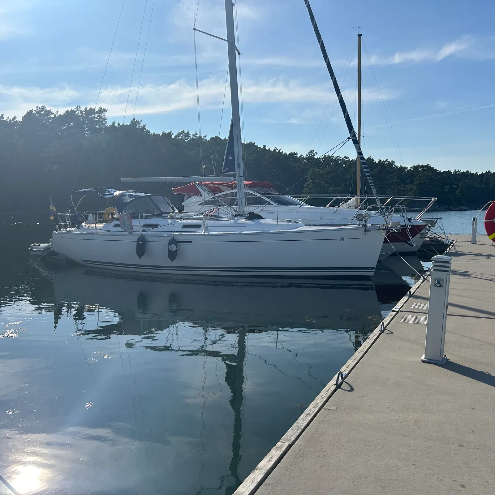 Segelbåtar och motorbåtar förtöjda vid en betongbrygga i en gästhamn i skärgården en solig sommarkväll, med stilla vatten och skog i bakgrunden.