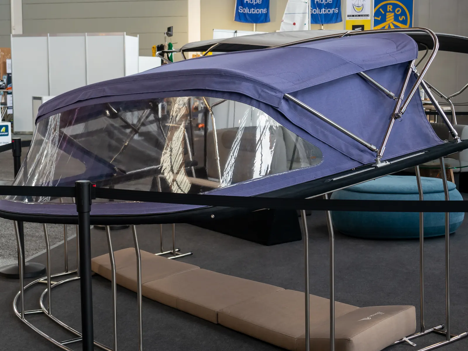 En blå sprayhood med bimini i textil på rostfria bågar, med klara plastfönster och beige dynor under, utställd i en mässhall.